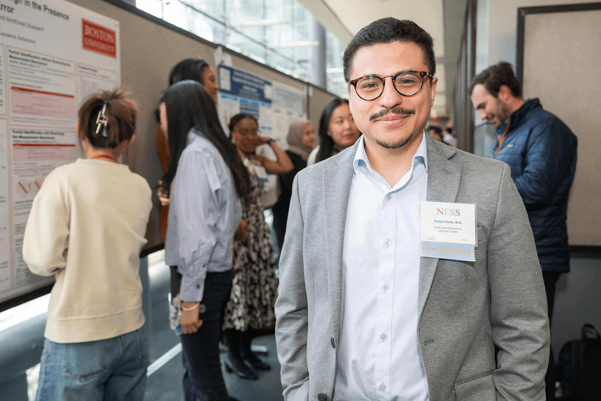 a ness judge standing and smiling in front of camer while people talk and look at poster presentations behind him