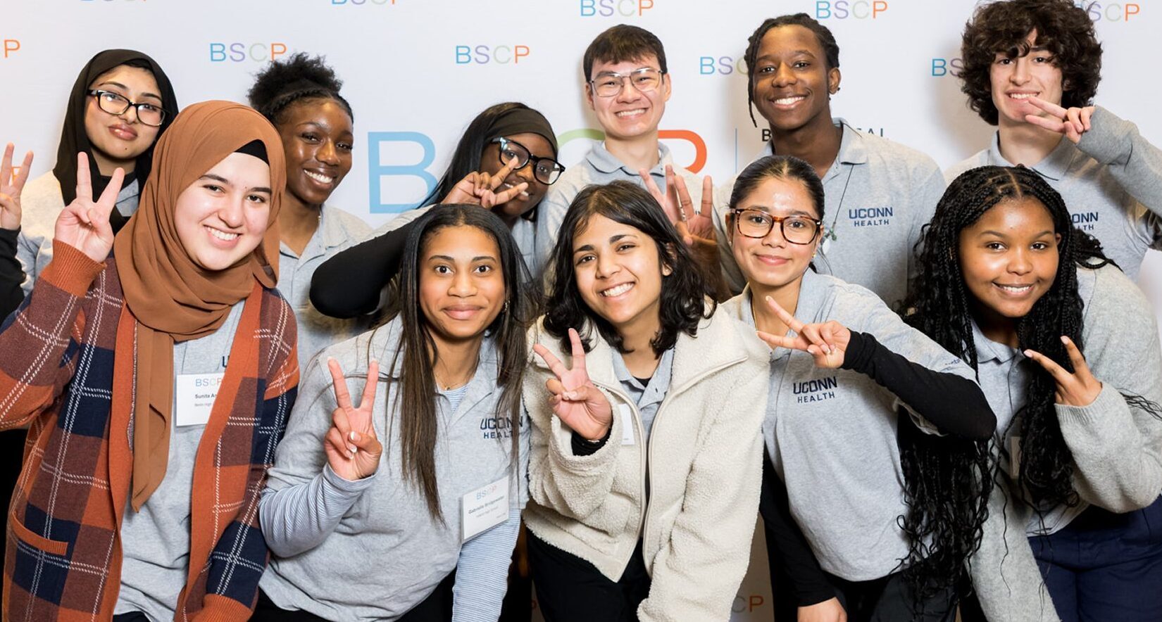 group of students standing in front of bscp step and repeat smiling and raising peace sign with fingers